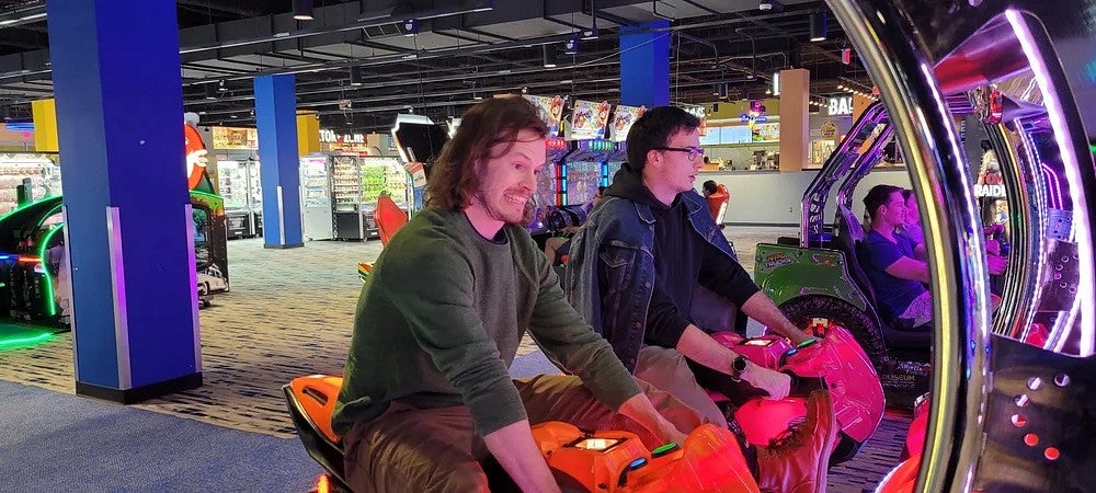 two men playing arcade games, where they ride on motorcycles in front of a big screen
