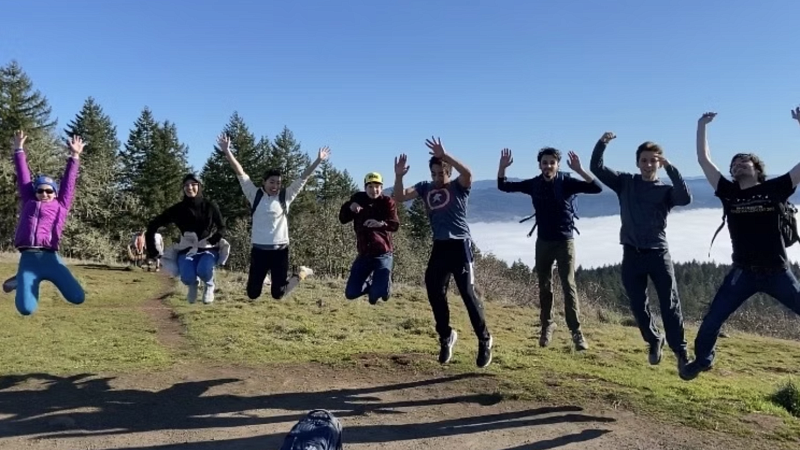 group of people jumping with their arms up, on top of Mt. pisgah - with trees and clouds in the background