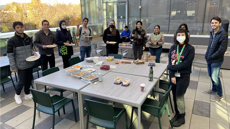 A group of people standing around the table, which has a variety of baked goods.