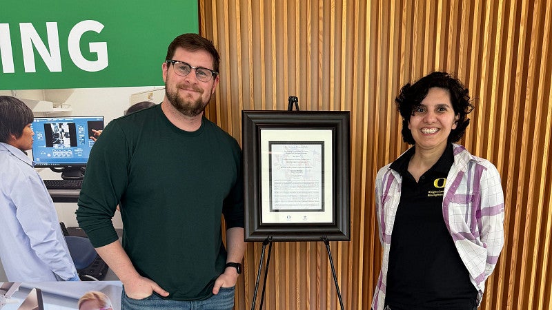 two people standing in front of a framed plaque, with a banner behind them