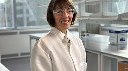 Woman in lab coat in lab with lab safety glasses smiling