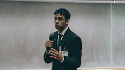 Student in formal attire with microphone in mid speech with chalkboard behind him
