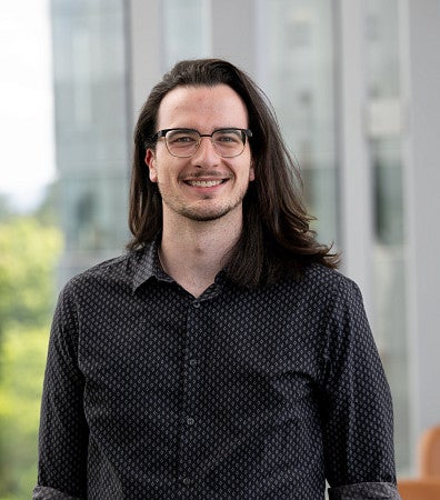 A man with long, black hair, and glasses, smiling for a headshot