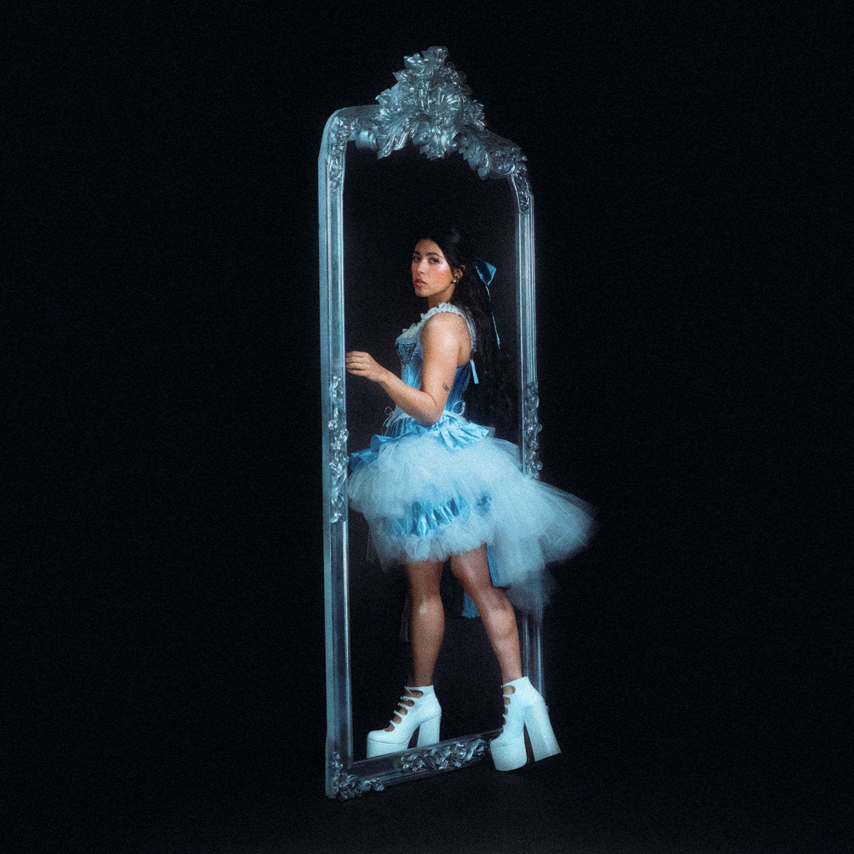 A black background with a woman in a blue dress and white boots walking into a mirror and turning to look at the camera 