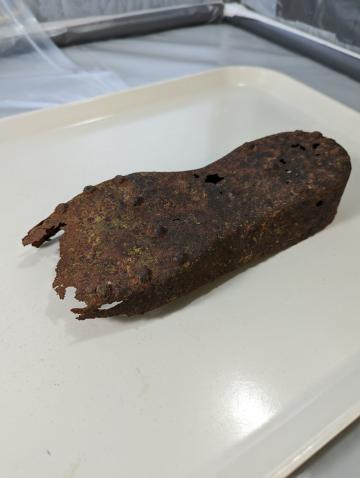 Rusty metal in the shape of an upside-down shoe lies on a white plastic tray. Thick spikes protrude from the top.