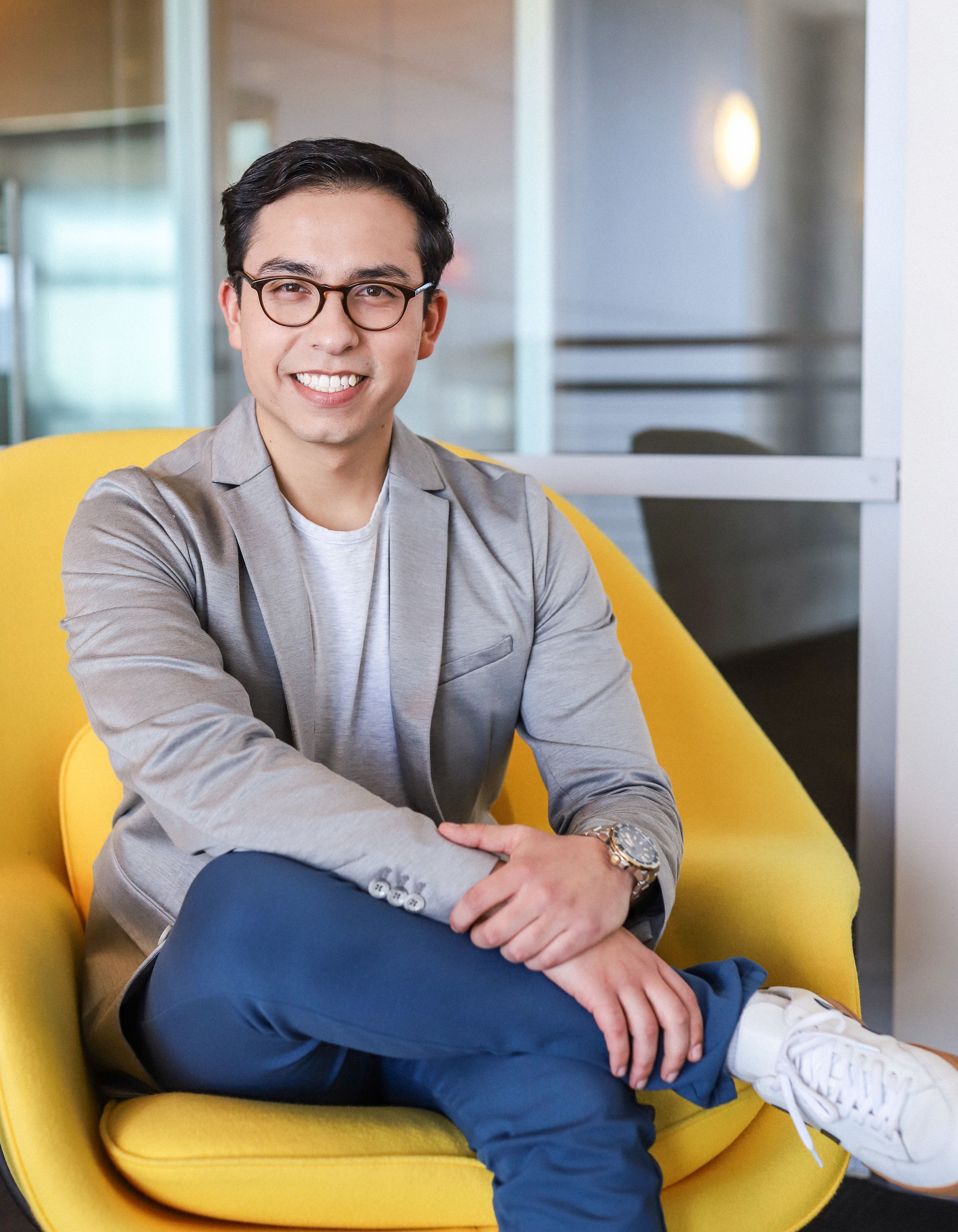Photo of Diego Rojas, sitting in a yellow chair with his arms resting on his knee, looking at the camera and smiling.