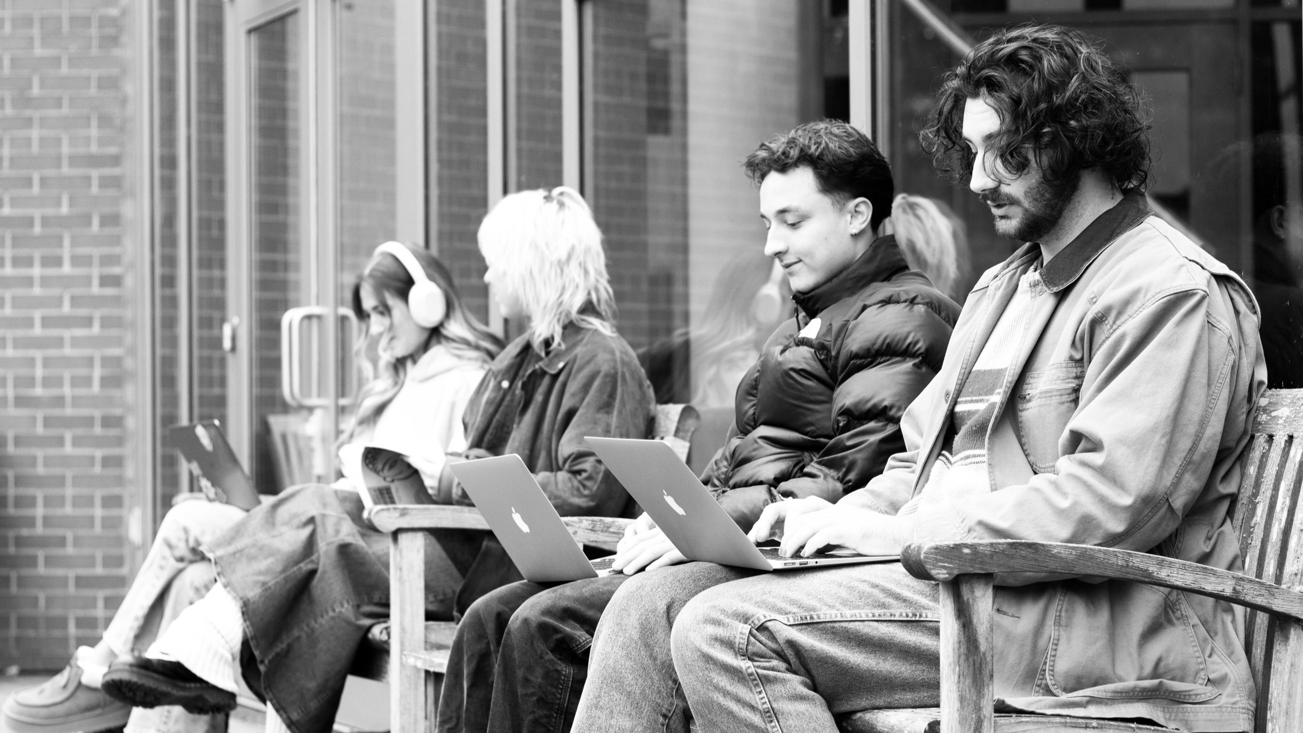 Four students sitting outside on benches, working on laptops.
