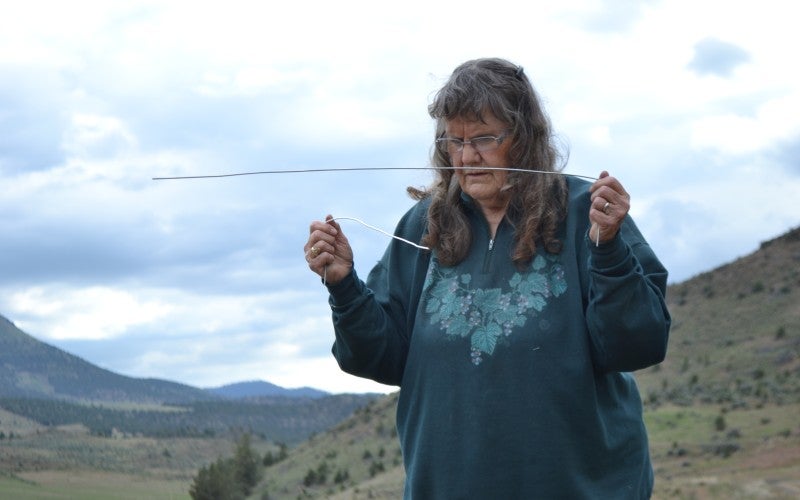 Betty stands outside wearing a green quarter zip jacket with leaf imagery and demonstrating her water witching technique.