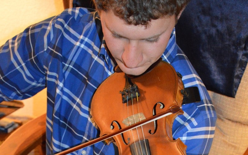 Eric sits in a chair playing a fiddle and wears a blue plaid shirt.