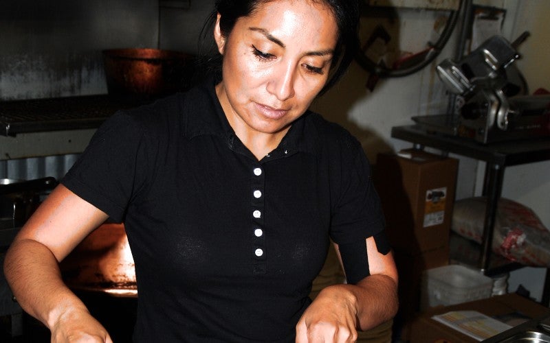 A woman stands in a kitchen and cuts cooked meat on a large white plate.