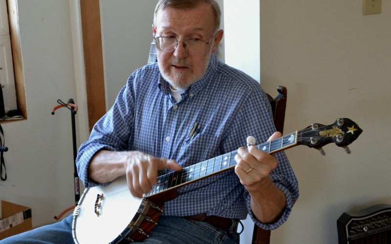 Hobe continues playing a banjo and wears a blue shirt.