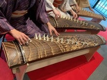 Three people sit playing kotos and wearing a different colored kimonos. One is purple, one is white, and one is yellow.