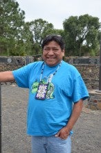 Thomas MorningOwl stands outside and gestures with his right hand. He is wearing a blue t-shirt and light blue jeans. He is also wearing a beaded necklace with a beaded pouch.