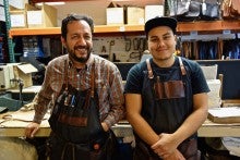 José Martinez stands in his workshop wearing a plaid shirt and an apron containing various tools.
