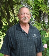 Dwight Cummins stands in front of trees. He wears a dark plaid polo that says "John Deere".