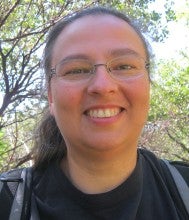 Patricia Whereat Phillips stands outside with trees in the background. She wears a black shirt and a gray backpack.