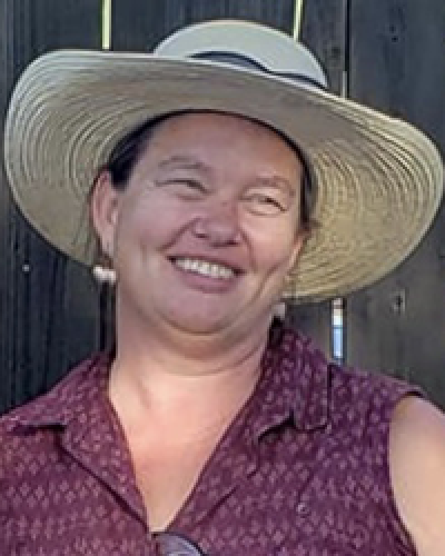 Correy McAtee stands in front of a tall wooden fence. She wears a beige sun hat and a dark purple tank top.