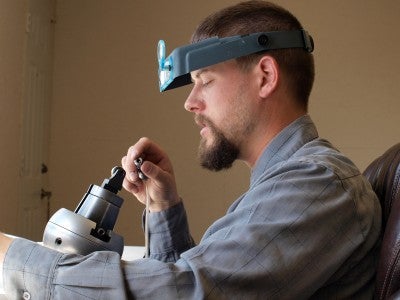 Pat Horlacher sits in front of smithing tools and examines a small silver piece. He wears a gray long sleeved shirt and a headpiece with a magnifying glass.