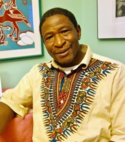 Baba Wagué Diakité sits in front of a mint green wall with two framed pictures. He wears a cream shirt with colorful embroidery.