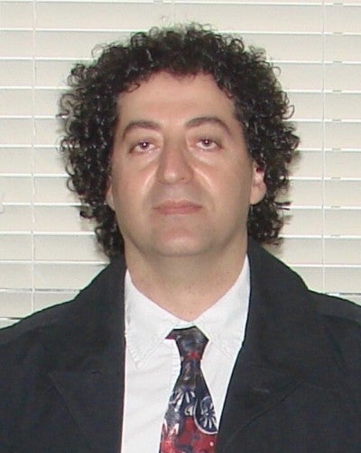 Houshang Sedighi stands against a white background. He wears a black suit and a red and black tie.