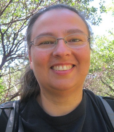 Patricia Whereat Phillips stands outside with trees in the background. She wears a black shirt and a gray backpack.