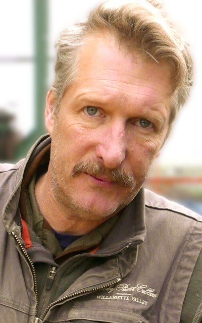 Rick DeFerrari stands outside and leans against a table. He wears a dark gray vest over a dark gray long sleeved shirt.