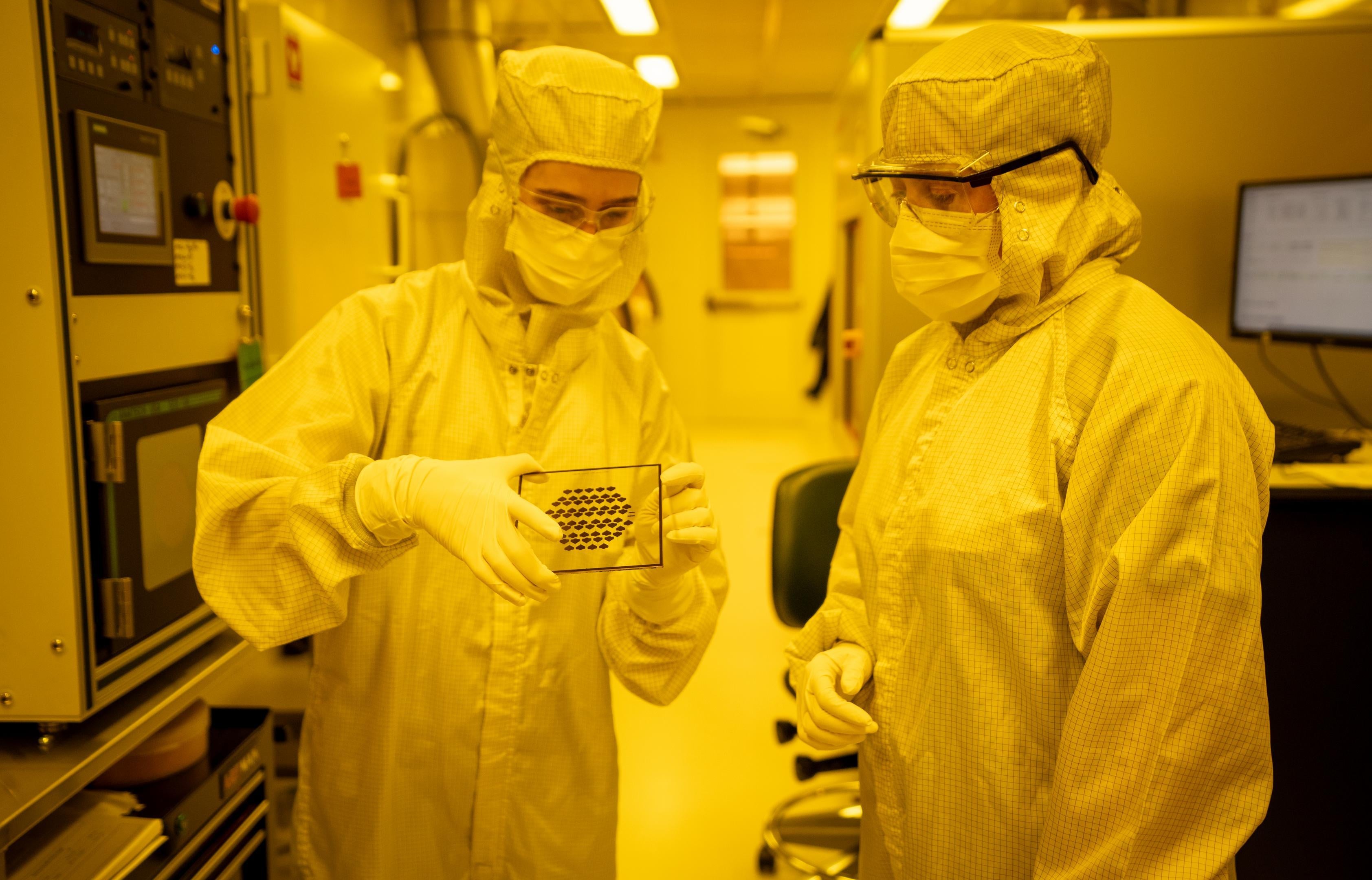 Two people wearing protective clothing examine a transparent slide with black dots on it.