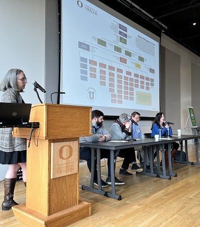 Person at podium and panel of four speakers in front of screen