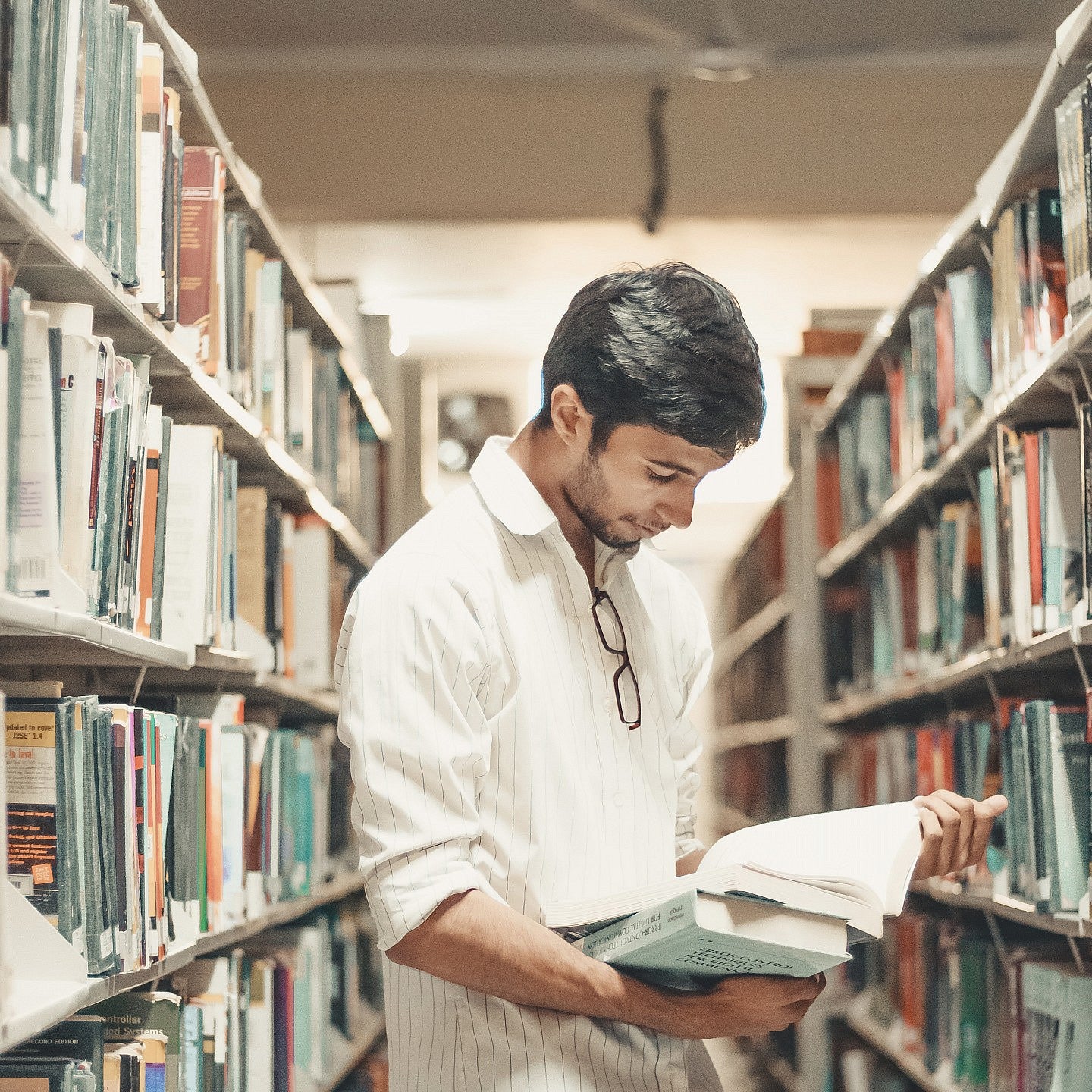 A male student perusing a book in the aisle between library bookshelves.