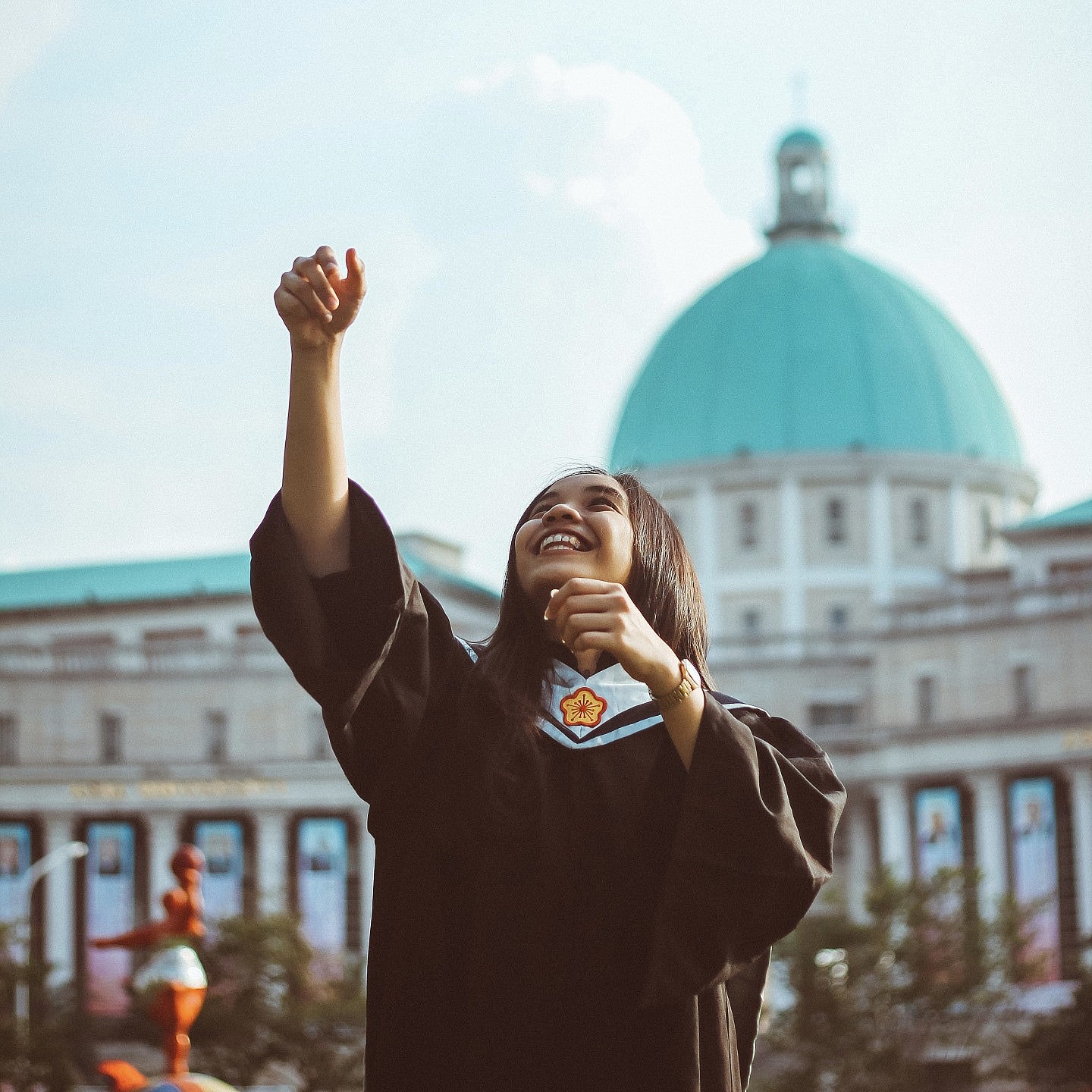 A female student in Graduation regalia looking & reaching up at the sky.