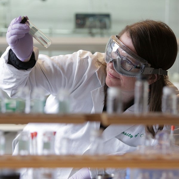 Undergraduate Researcher wearing goggles and a lab coat looking at a test tube.