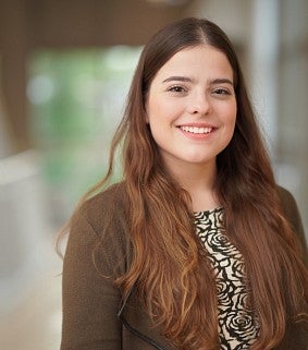 Woman with long brown hair wearing a brown jacket and black and white top smiles at the camera