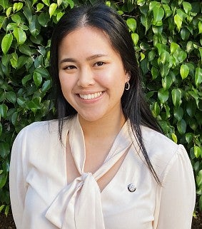 Woman with long dark hair smiling; she wears a white shirt and purple skirt.