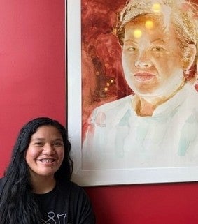 Young woman with long dark hair stands next to a portrait
