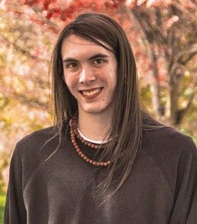 Person with long brown hair and brown shirt smiles at the camera; autumn leaves and trees in the background