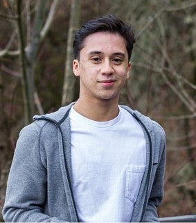 Young man smiling; he has short brown hair, white T-shirt, and grey sweatshirt; in the background are trees