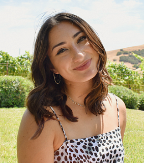 Woman with brown hair, hoop earrings, and a blue-and-white spotted shirt smiles in front of green hills and trees and a blue sky with clouds