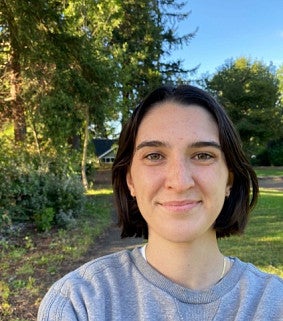 Young woman with short brown hair and wearing a gray sweatshirt smiles in front of green trees and grass and a blue sky