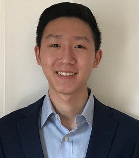 Young man with short black hair wearing a light blue button-down shirt and dark blue jacket smiles at the camera in front of a white background