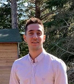 Young man with short brown hair, wearing a pink collared shirt, smiles at the camera in front of a brown and green building and green trees