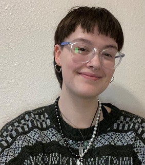 Woman with short brown hair, glasses, heart-shaped ear-rings, a grey patterned sweater, and ying-yang necklace, smiles in front of a white background