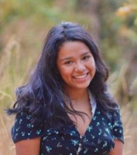 Woman with long dark hair and a dark blue shirt smile at the camera; trees are blurry in the background