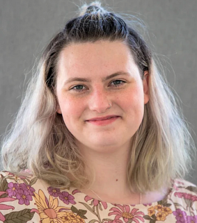 Woman with shoulder-length blonde and dark hair and wearing a floral pink and yellow shirt smiles in front of a light grey background