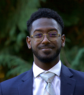 Man with short black hair and glasses, a cropped beard and moustache, wearing a blue suit and light grey tie smiles; green trees in the background