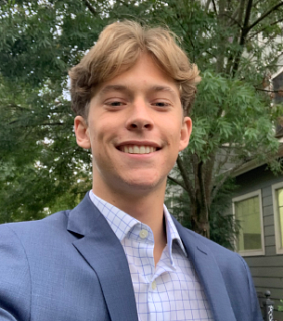 Young white man with light brown hair parted in the middle of his head; wearing a light blue jacket and collared shirt; trees and a building behind him
