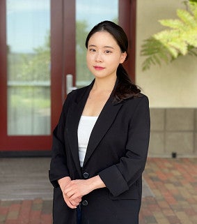 Young Asian woman with long black hair pulled back, holding her hands in front of her; wearing a black jacket and white shirt; glass-wood doors behind her
