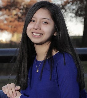Young woman with long dark hair, seated, hands crossed in front of her and resting on her knee; long-sleeved purple shirt; autumn leaves behind her