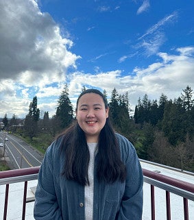Young Asian woman with long dark hair; wearing a blue long-sleeved top and white shirt; blue skies, clouds, a road and rooftop behind her
