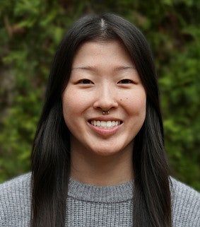 Asian woman with long black hair and grey jersey in front of greenery smiles at camera
