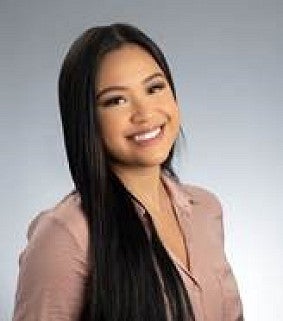 Young Asian woman with long dark hair tilts her head and smiles at the camera; wearing a light brown shirt; white-grey wall behind her